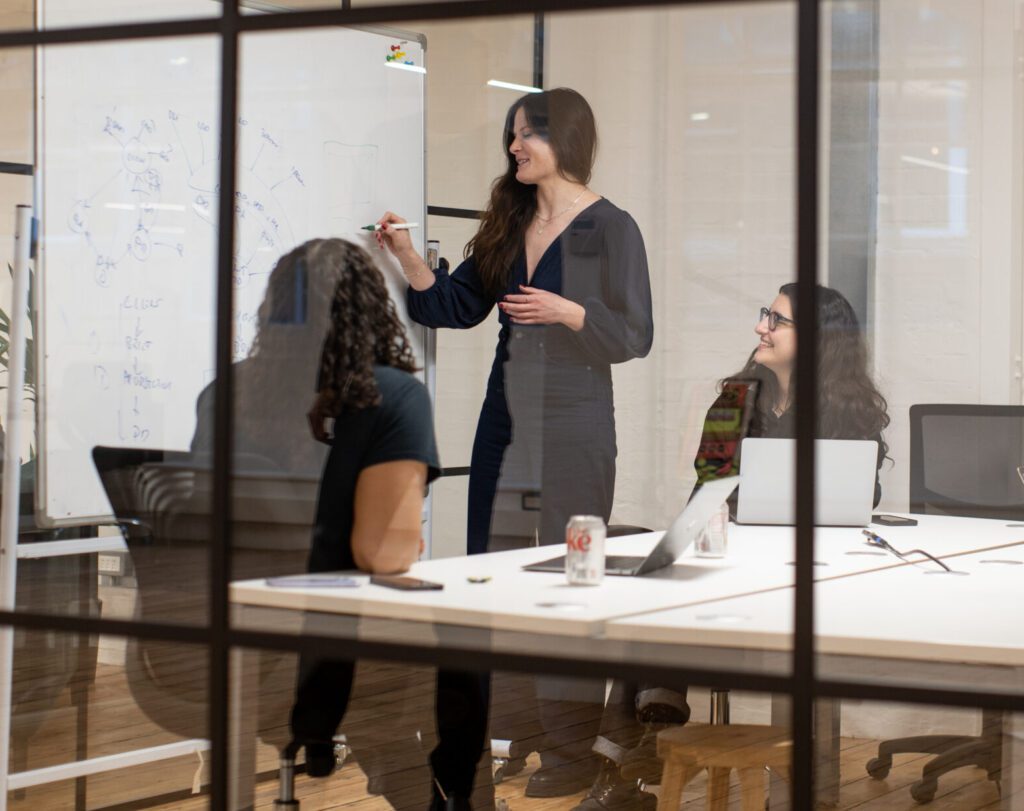 Picture through a meeting room glass wall with a group of people discussing a white board presentation